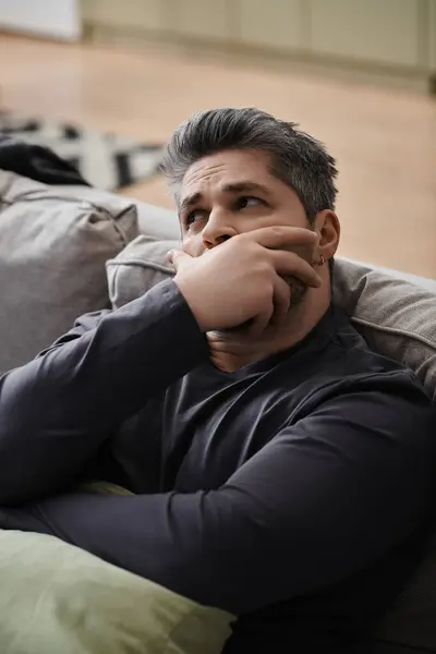 A handsome man lost in thought, resting on a couch in a sunlit living room with a serene atmosphere. — Stock Photo