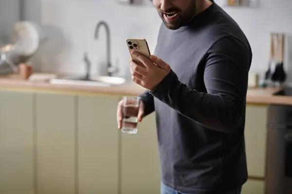 A dapper man smiles at his smartphone, holding a glass of water in a stylish kitchen setting. — Stock Photo