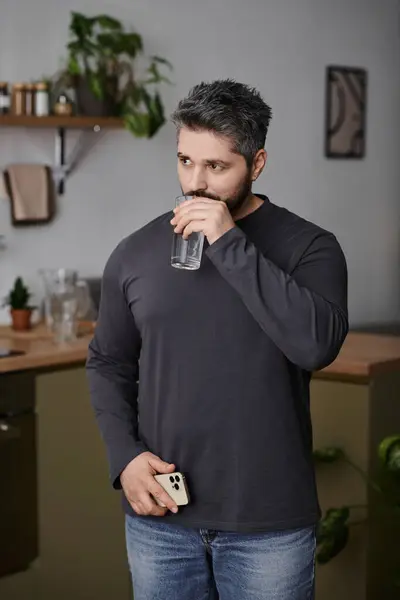 Um homem elegante leva um momento para beber água, absorvida pelo pensamento, em um ambiente de cozinha quente. — Fotografia de Stock