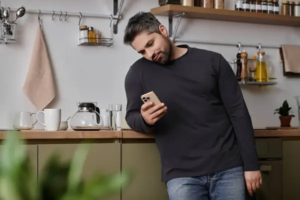 Bello uomo si appoggia contro un bancone della cucina, assorbito nel suo smartphone sorseggiando caffè. — Foto stock