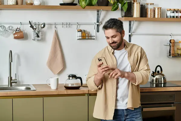 Bonito homem gosta de seu tempo em uma cozinha moderna, percorrendo seu smartphone com um sorriso. — Fotografia de Stock