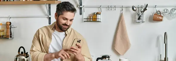 A stylish man with a cheerful expression checks his smartphone in a modern kitchen setting. — Stock Photo