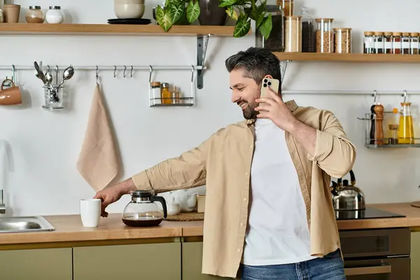 Un hombre alegre vierte café y chats en el teléfono en una cocina acogedora. - foto de stock