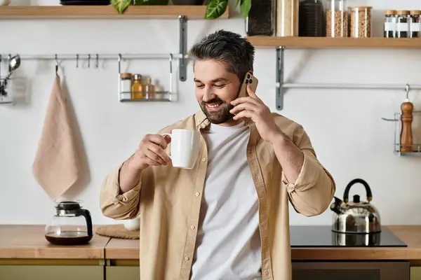 Un hombre con una sonrisa encantadora bebe café y charla por teléfono en una cocina moderna. — Stock Photo