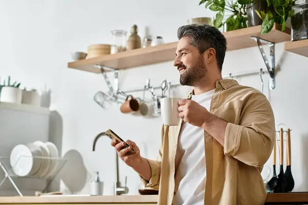 Bright kitchen filled with plants where a handsome man savors coffee while glancing at his phone. — Stock Photo