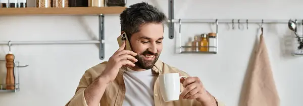 Очаровательный мужчина улыбается, потягивая кофе и разговаривая по телефону на современной кухне. — стоковое фото