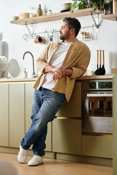 Un hombre guapo se apoya casualmente contra un mostrador de cocina elegante mientras se pierde en el pensamiento. - foto de stock