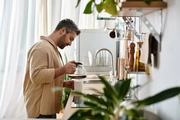 Un hombre guapo vierte café caliente en una taza en medio de plantas de cocina y la luz del sol. - foto de stock
