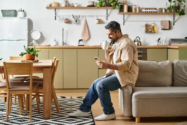Stylish man relaxes in a modern kitchen, sipping coffee and scrolling through messages on his phone. — Stock Photo