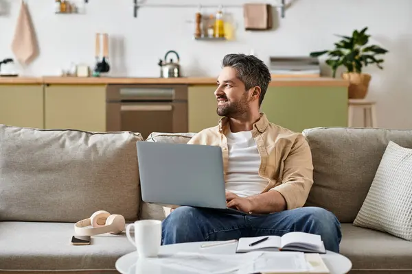 Un bell'uomo si siede comodamente su un divano, concentrandosi sul suo computer portatile in un elegante soggiorno. — Foto stock