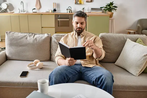 A handsome man enjoys a quiet moment reading a book on a comfortable sofa in his home. — Stock Photo