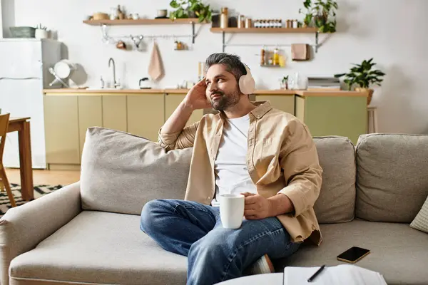 Hombre guapo con auriculares se sienta en un sofá, saboreando una bebida caliente en una cocina elegante. - foto de stock
