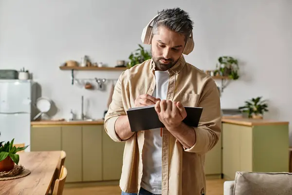 Un bell'uomo che indossa le cuffie si concentra sul prendere appunti in una cucina elegante con piante.. — Foto stock