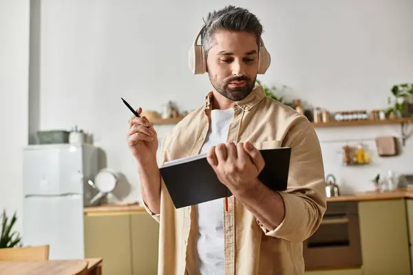 Un bell'uomo con le cuffie scrive in un quaderno, circondato da una calda atmosfera da cucina. — Foto stock