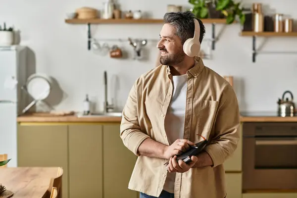 Un hombre alegre escucha música mientras sostiene un dispositivo, sonriendo en una cocina elegante. - foto de stock