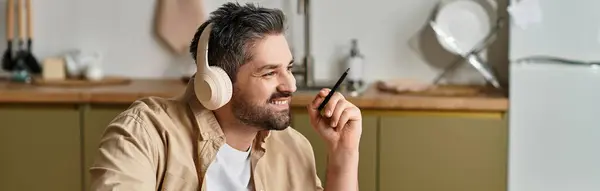 Un hombre alegre con auriculares sonríe mientras escribe ideas en una acogedora cocina. - foto de stock