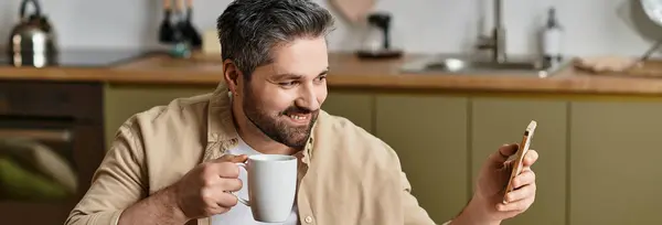 Smiling man sips coffee while looking at his smartphone in a bright kitchen setting. — Stock Photo