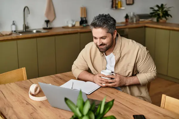 A handsome man smiles as he sips coffee and writes notes at a wooden table in a modern kitchen. — Stock Photo
