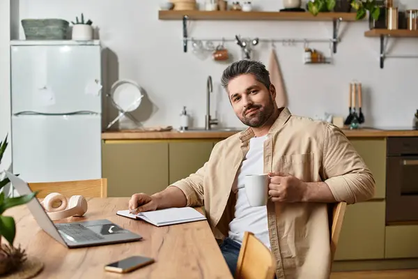 A man sits comfortably at a wooden table in a modern kitchen, enjoying a warm drink and working. — Stock Photo