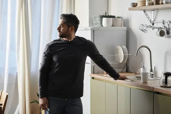 A handsome man stands thoughtfully in a cozy kitchen, reflecting by the window during midday. — Stock Photo