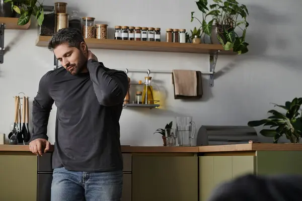 Un bel homme se tient dans une cuisine chic, perdu dans la pensée avec sa main sur le cou. — Photo de stock