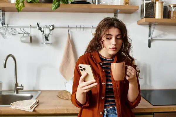 A woman sits in her kitchen, holding a warm cup and gazing at her phone, deep in thought. — Stock Photo
