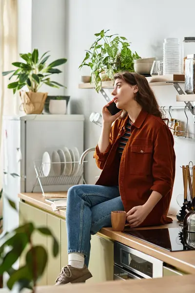 In una cucina illuminata dal sole, una donna siede sul bancone, parlando al telefono con un'espressione distante. — Foto stock