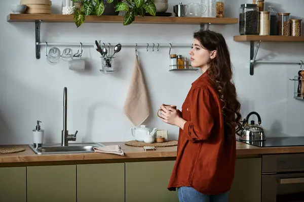 Une femme se tient pensivement dans une cuisine chaude, tenant une tasse et perdue dans ses pensées. — Stock Photo