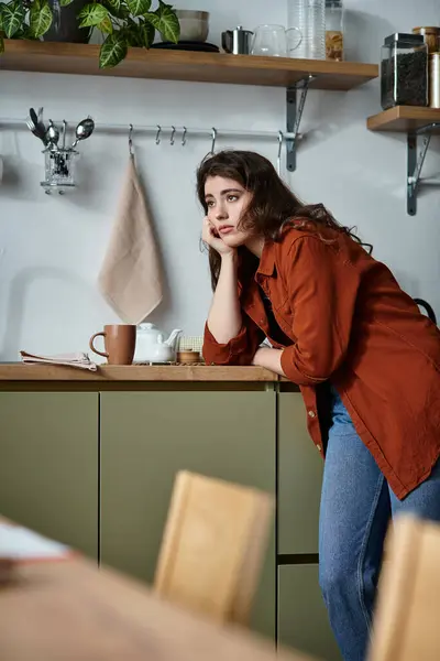 Una giovane donna si appoggia su un bancone della cucina, persa nel pensiero in un ambiente tranquillo del mattino. — Foto stock
