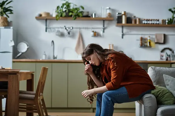 A woman sits on a couch in a cozy kitchen, visibly troubled and lost in thought as shadows fall. — Stock Photo