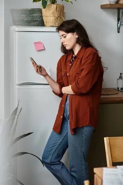 A woman stands pensively by a refrigerator, lost in thought while holding her smartphone. — Stock Photo