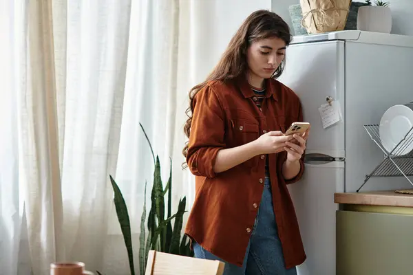 A woman stands in a serene kitchen, absorbed in her phone, reflecting her inner struggles. — Stock Photo