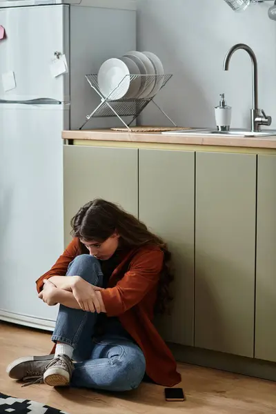 Une femme assise seule sur le sol de la cuisine, embrassant ses genoux, perdue dans ses pensées et ses émotions. — Stock Photo
