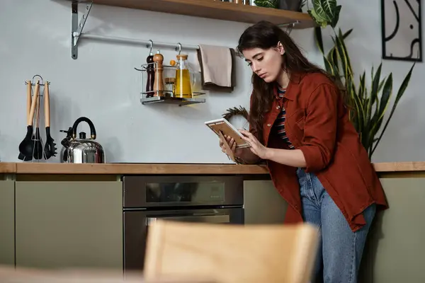 Dans une cuisine chaude, une femme se penche contre le comptoir, profondément dans la pensée alors qu'elle examine son carnet. — Photo de stock