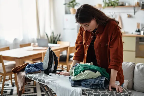 Eine Frau steht an einem Bügelbrett, überwältigt von Wäsche, verloren in Gedanken in ihrem Haus. — Stockfoto