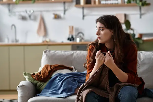 A woman sits on a couch with a pensive expression, holding her sweater tightly as she reflects. — Stock Photo