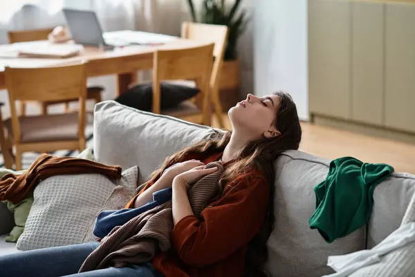 Una donna si siede su un divano, sopraffatta dalle emozioni in mezzo a un mucchio di vestiti, in cerca di conforto. — Foto stock