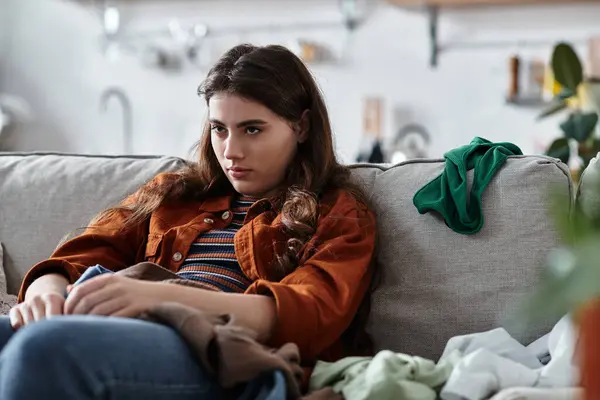 Una donna si siede tranquillamente su un divano, circondata da vestiti disordinati, mostrando la sua tristezza interiore.. — Foto stock