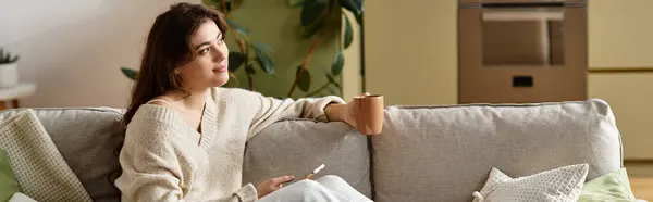 In a serene living room, a woman sits comfortably on the couch, lost in thought and holding a mug. — Stock Photo