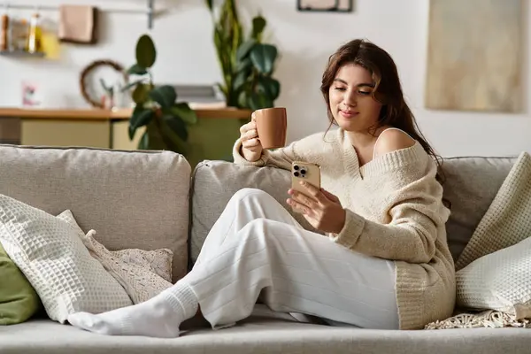 A young woman sits on a couch with a warm drink, absorbed in her phone and deep in thought. — Stock Photo