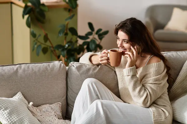 A woman sits on a cozy couch in a softly lit room, sipping a warm drink and lost in thought. — Stock Photo