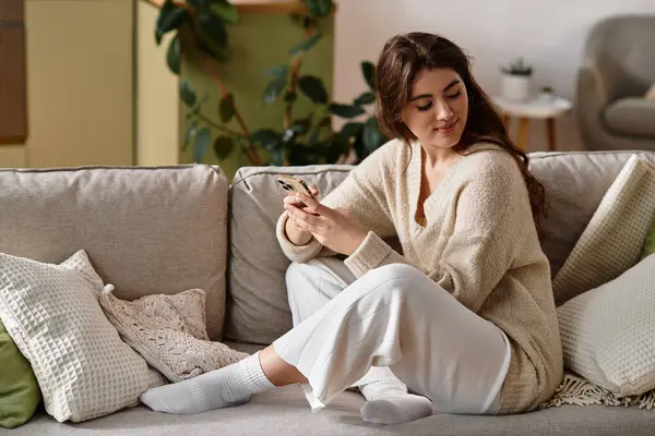 A young woman sits on a sofa, lost in thought, reflecting on her feelings while using her phone. — Stock Photo