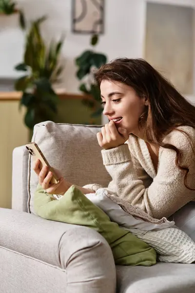 A woman sits on a couch, immersed in her thoughts and the glow of her phone screen. — Stock Photo