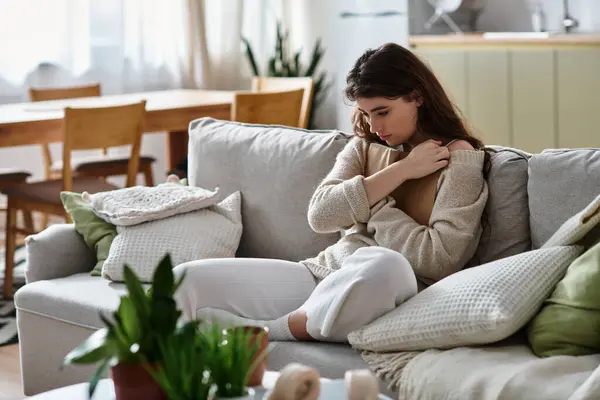 Una donna vestita con un morbido maglione si siede su un divano, persa nel pensiero mentre circondata da un arredamento morbido. — Foto stock