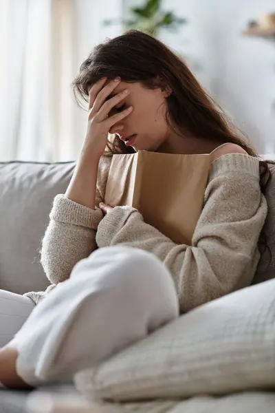 Eine Frau sitzt auf einem Sofa und klammert sich an ein Buch, während sie in einem sanft beleuchteten Raum von ihren Emotionen überwältigt wird. — Stockfoto