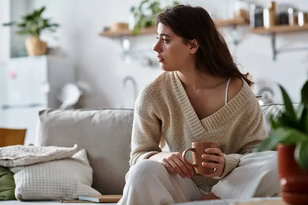 Une femme assise sur un canapé, tenant une boisson chaude, perdue dans la pensée alors qu'elle est entourée d'un décor confortable. — Stock Photo