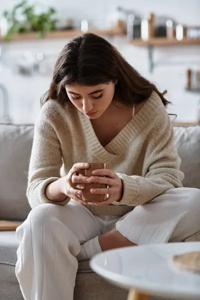 Sitting on a cozy couch, a woman holds a cup in her hands, lost in contemplative thoughts. — Stock Photo