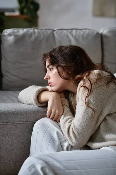 A woman sits on a couch, lost in thought and grappling with deep emotions as she looks away. — Stock Photo