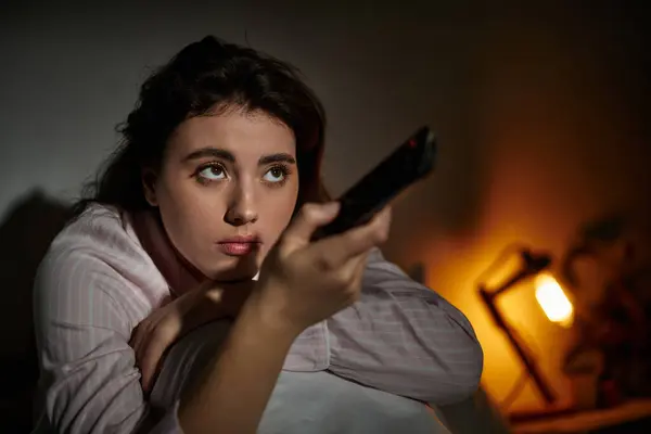 A woman sits alone in her dimly lit room, lost in thought while holding a remote. — Stock Photo