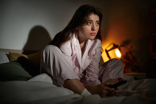 A woman sits quietly on her bed, lost in deep thought, surrounded by shadows and a warm glow. — Stock Photo